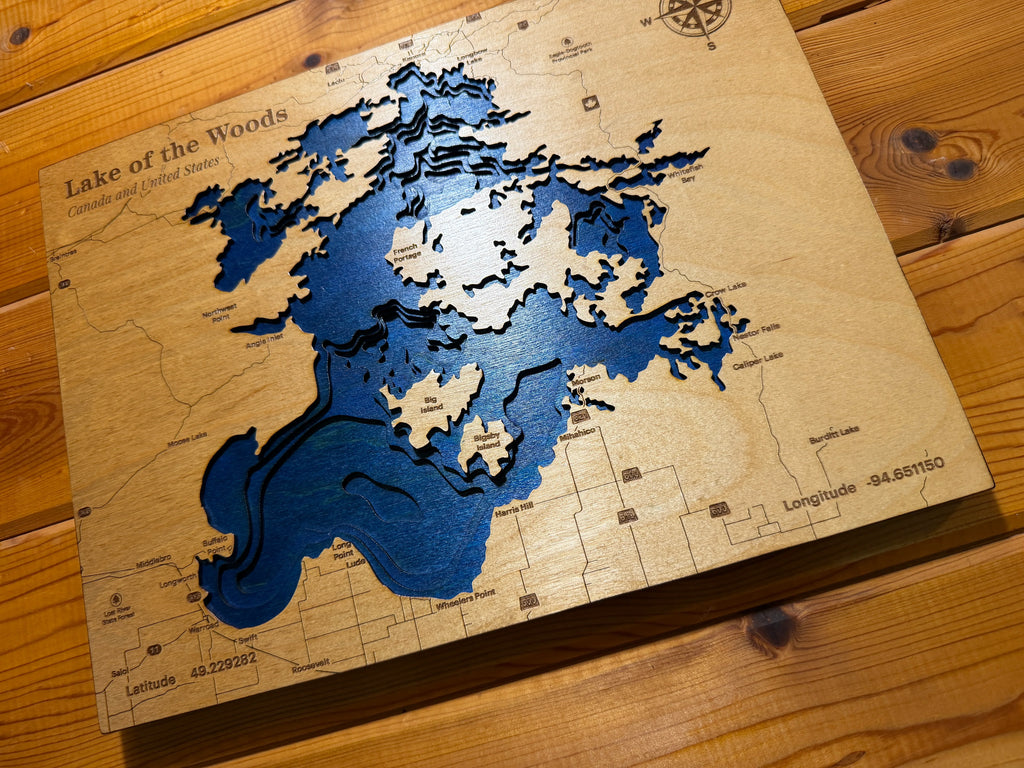 Layered wood map of Lake of the Woods, Canada and US, with blue depth layers and engraved landmarks, displayed on wooden surface.