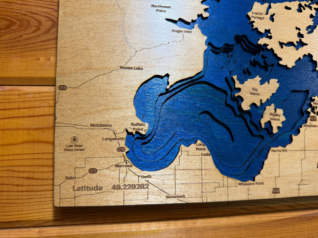 Layered wood lake map with blue water depths, carved place names, and latitude detail on natural wood background