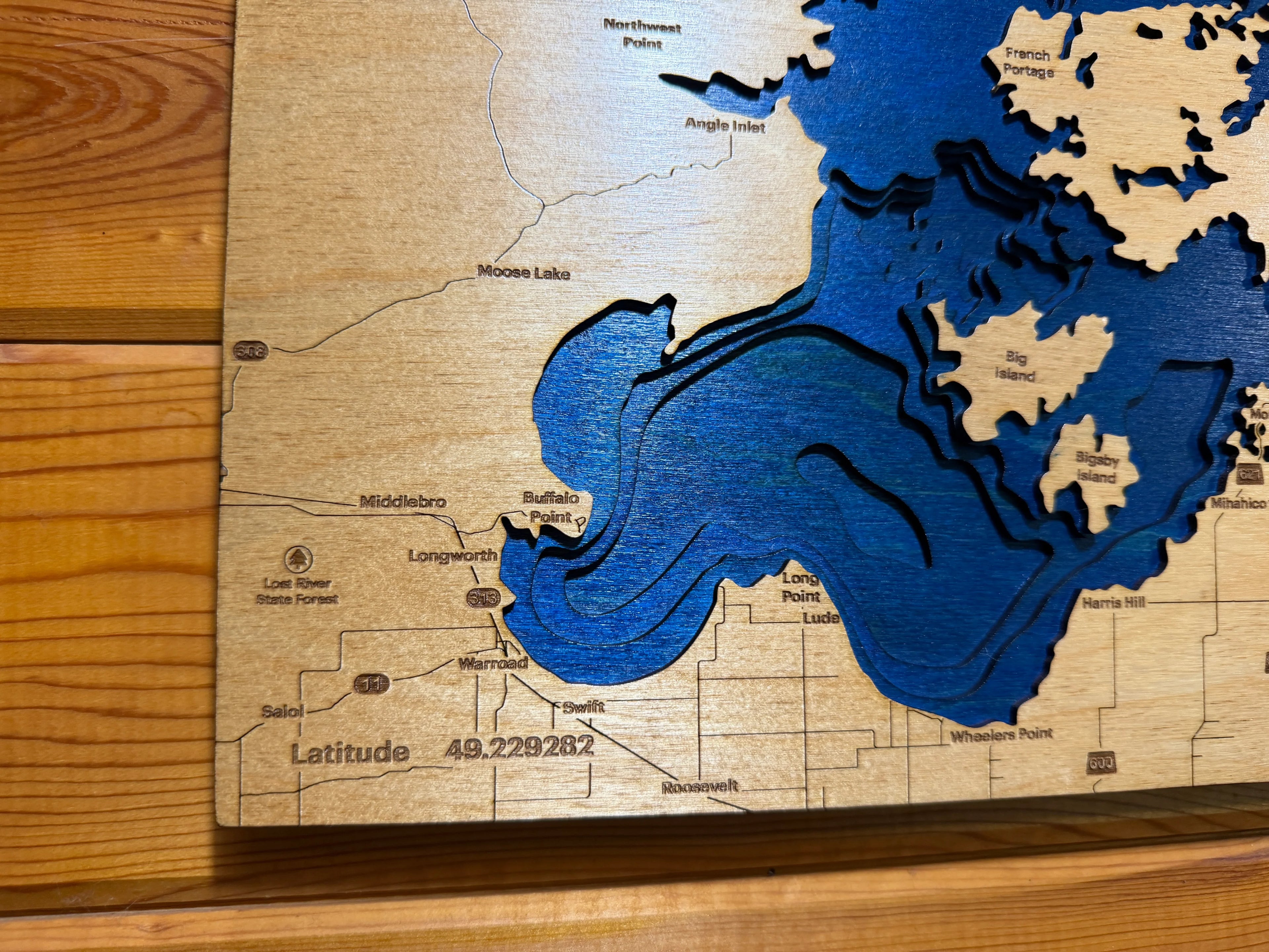 Layered wood lake map with blue water depths, carved place names, and latitude detail on natural wood background