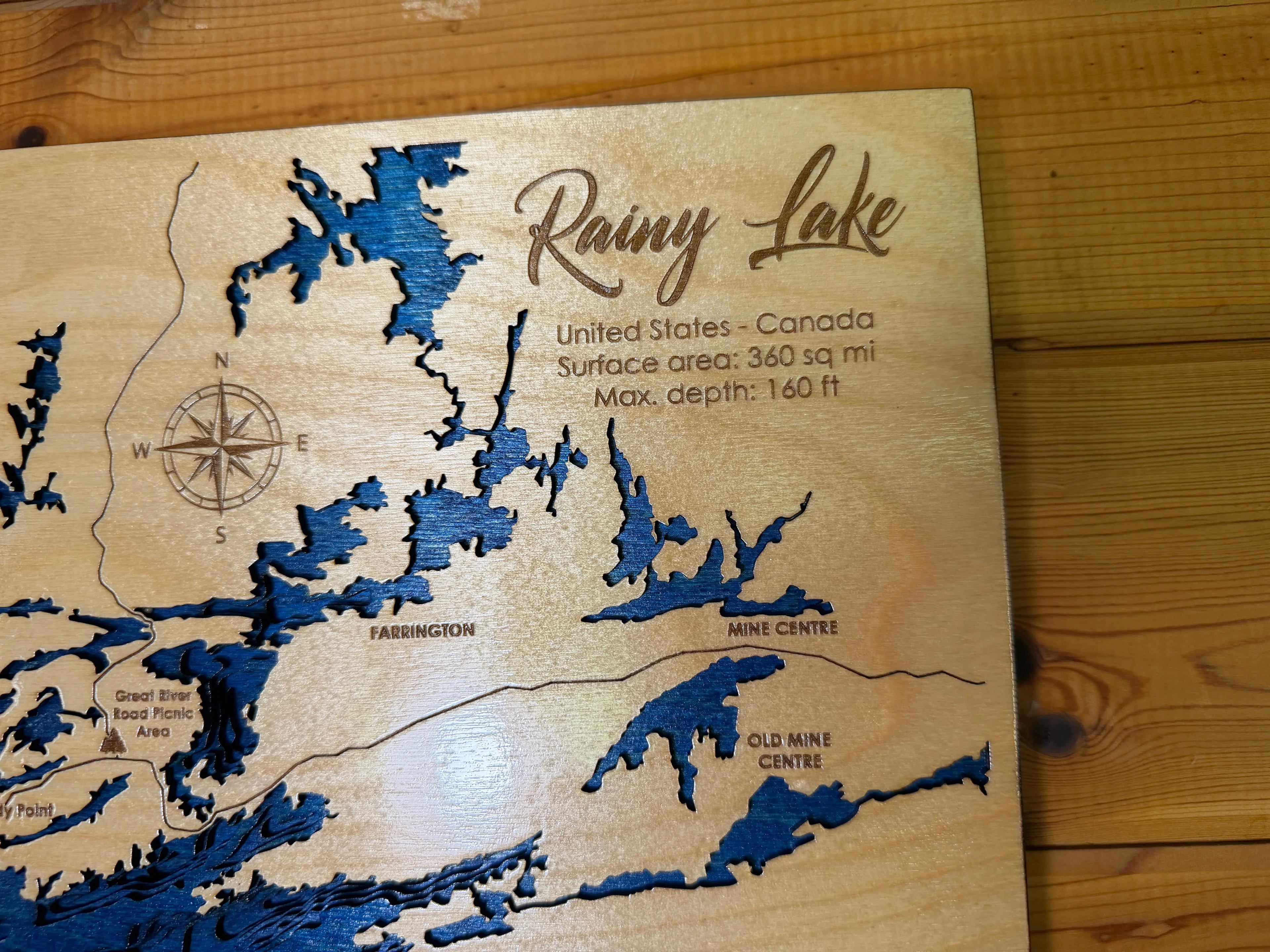 Layered wood map of Rainy Lake, USA-Canada border, with blue water cutouts and engraved details like Farrington and Mine Centre.