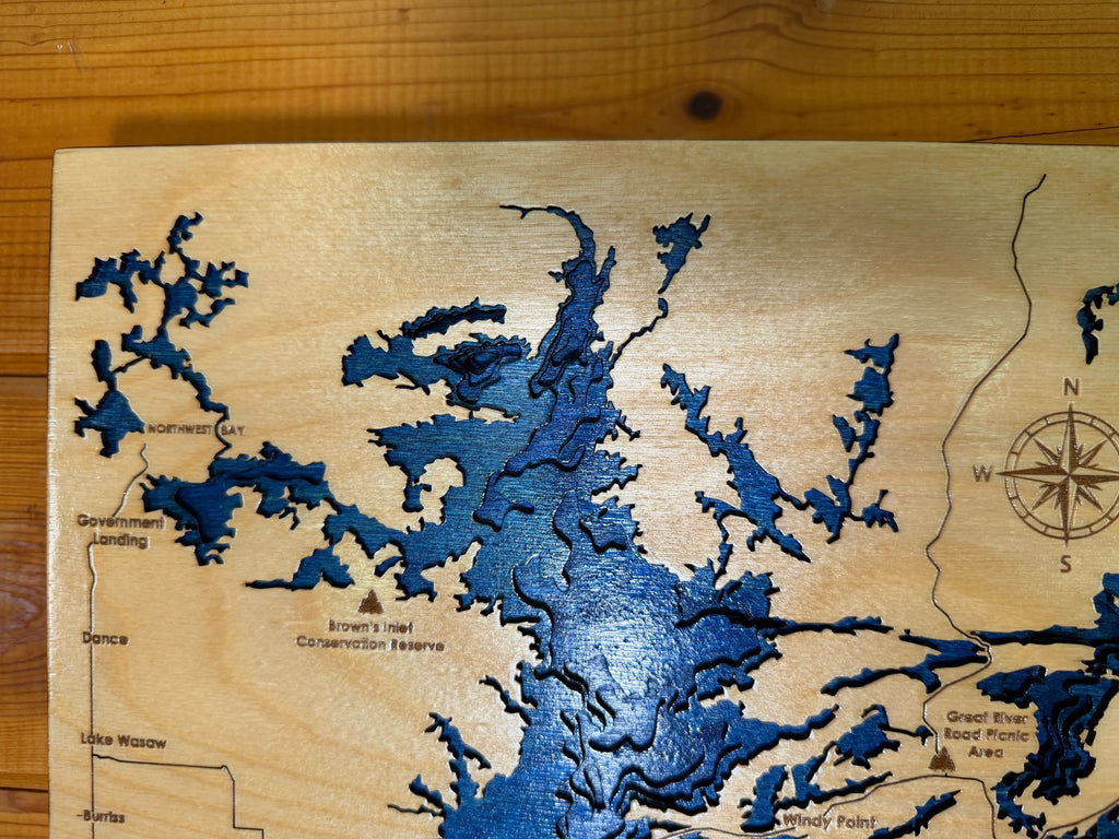 Layered wood lake map with blue depth layers, compass rose, and engraved locations like Northwest Bay and Lake Wasaw.