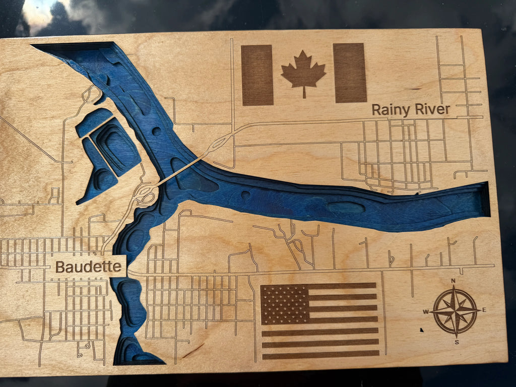 A rectangular wooden plaque featuring an engraved topographic-style map with a blue recessed river channel, labeled 'Rainy River' and 'Baudette.' The wood shows natural grain and includes a Canadian flag, a U.