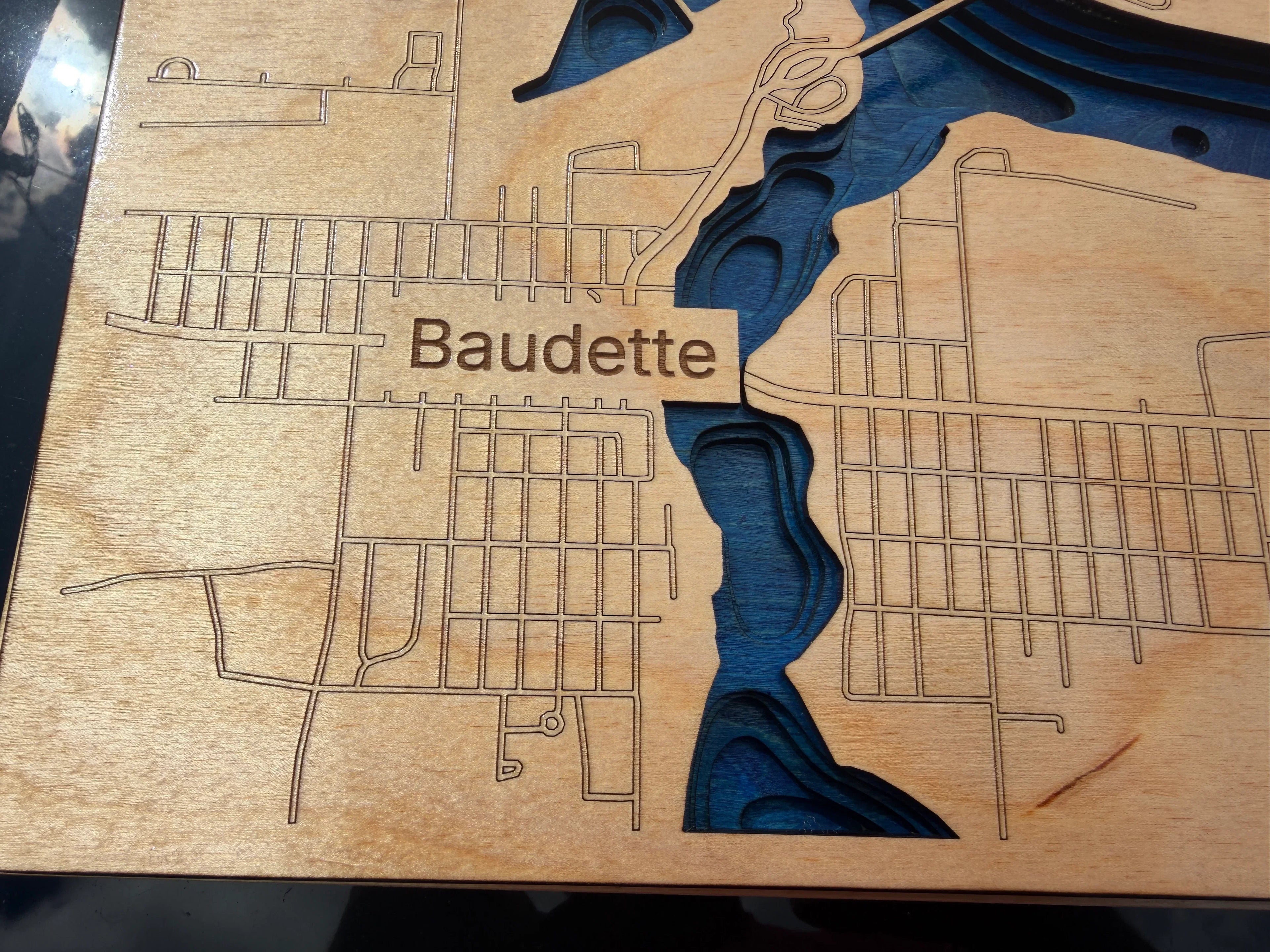 Close-up of a laser-engraved wooden map plaque featuring the name Baudette, with layered dark blue water channels carved into a light natural wood surface.