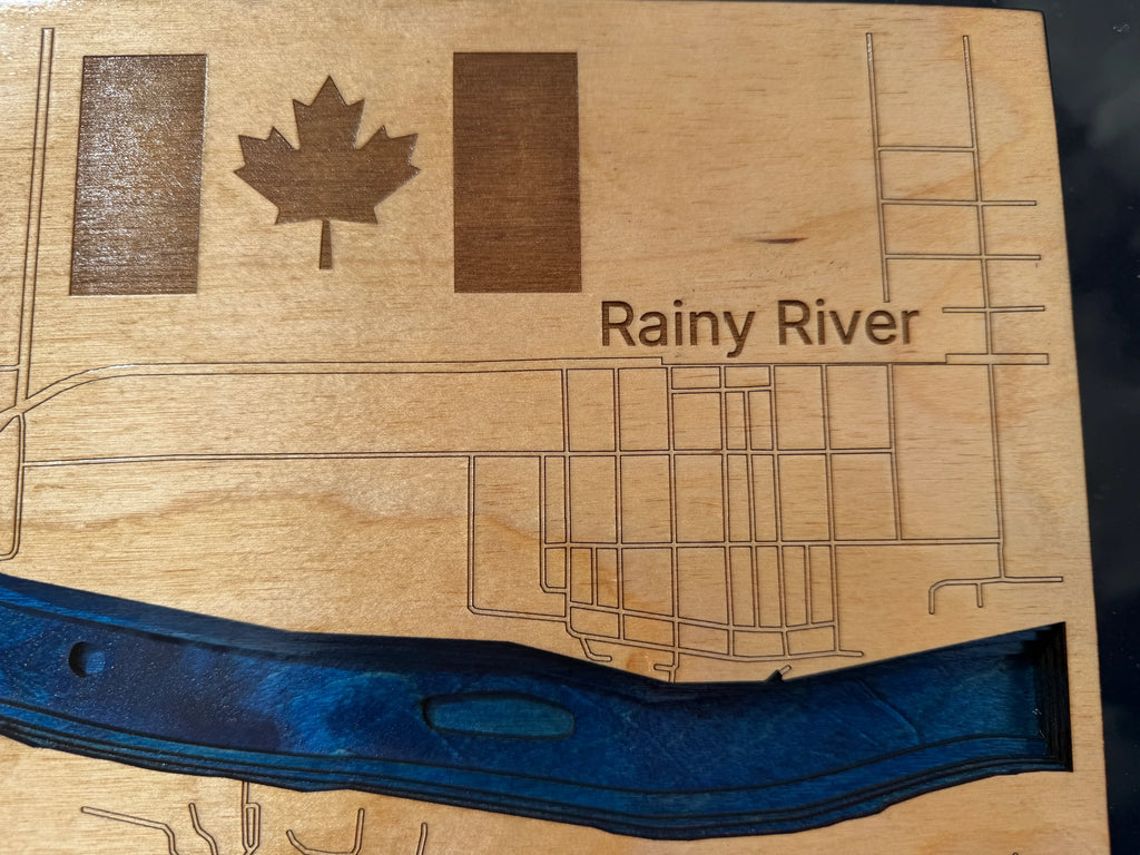 A close-up of a wooden plaque featuring an engraved map design labeled 'Rainy River,' with a maple leaf and two vertical rectangular shapes near the top, plus a dark blue recessed river element along the bottom.