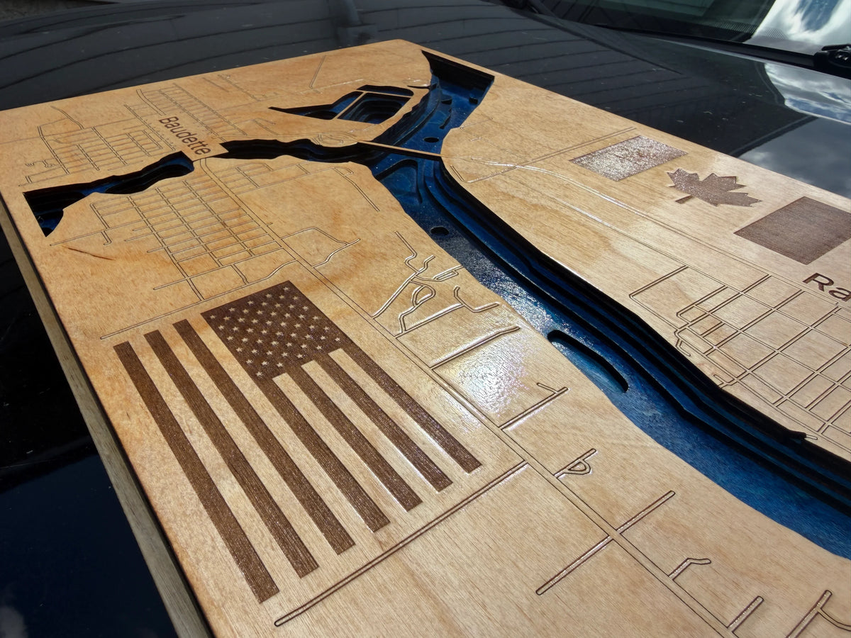A close-up of a wooden plaque or wall art piece featuring an engraved American flag, city street map lines, and a dark blue river-like channel running through the center, photographed outdoors on a dark surface.