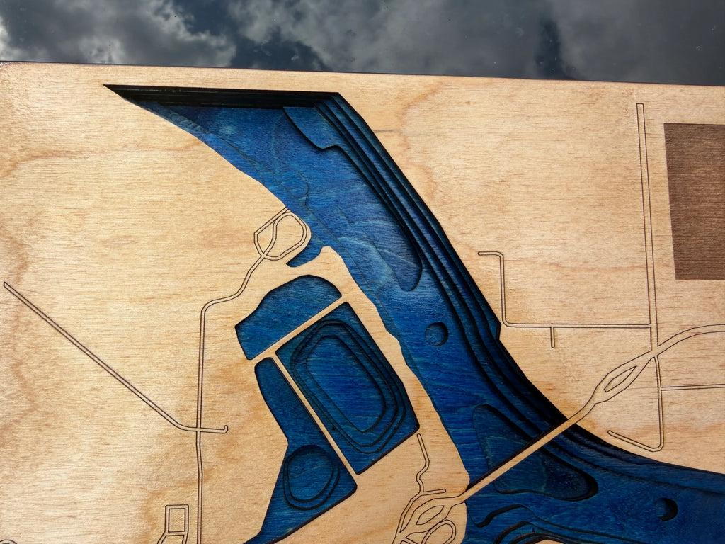 Close-up of a wooden map plaque featuring a stylized blue river or inlet carved into a light natural wood board, with engraved contour lines and street-like outlines.