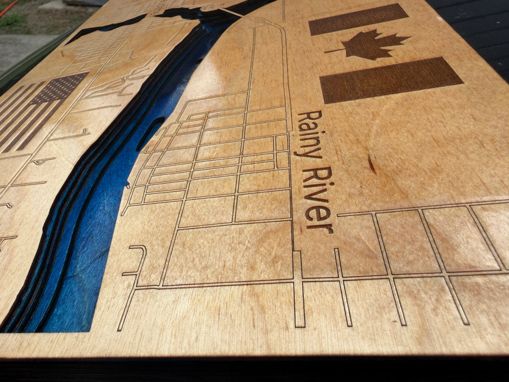 A close-up of a laser-engraved plywood wall art piece depicting the Rainy River, with a blue recessed river channel, street grid lines, Canadian and American flag motifs, and visible wood grain.