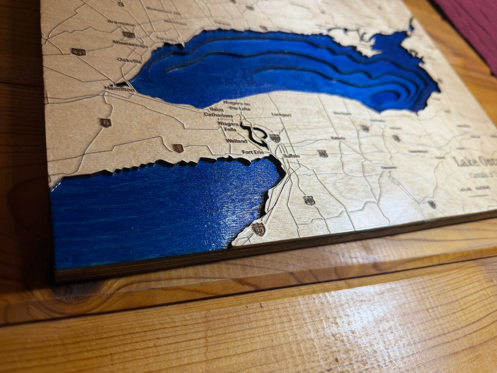 A close-up of a 3D wooden map plaque of Lake Ontario, featuring layered wood cutouts that form the blue lake shape against a natural wood grain background.