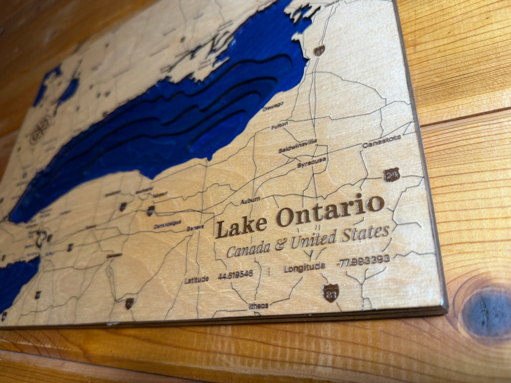 A close-up of a wooden map plaque featuring Lake Ontario and surrounding Canada and United States routes, with engraved text and dark blue water areas on a light wood background.