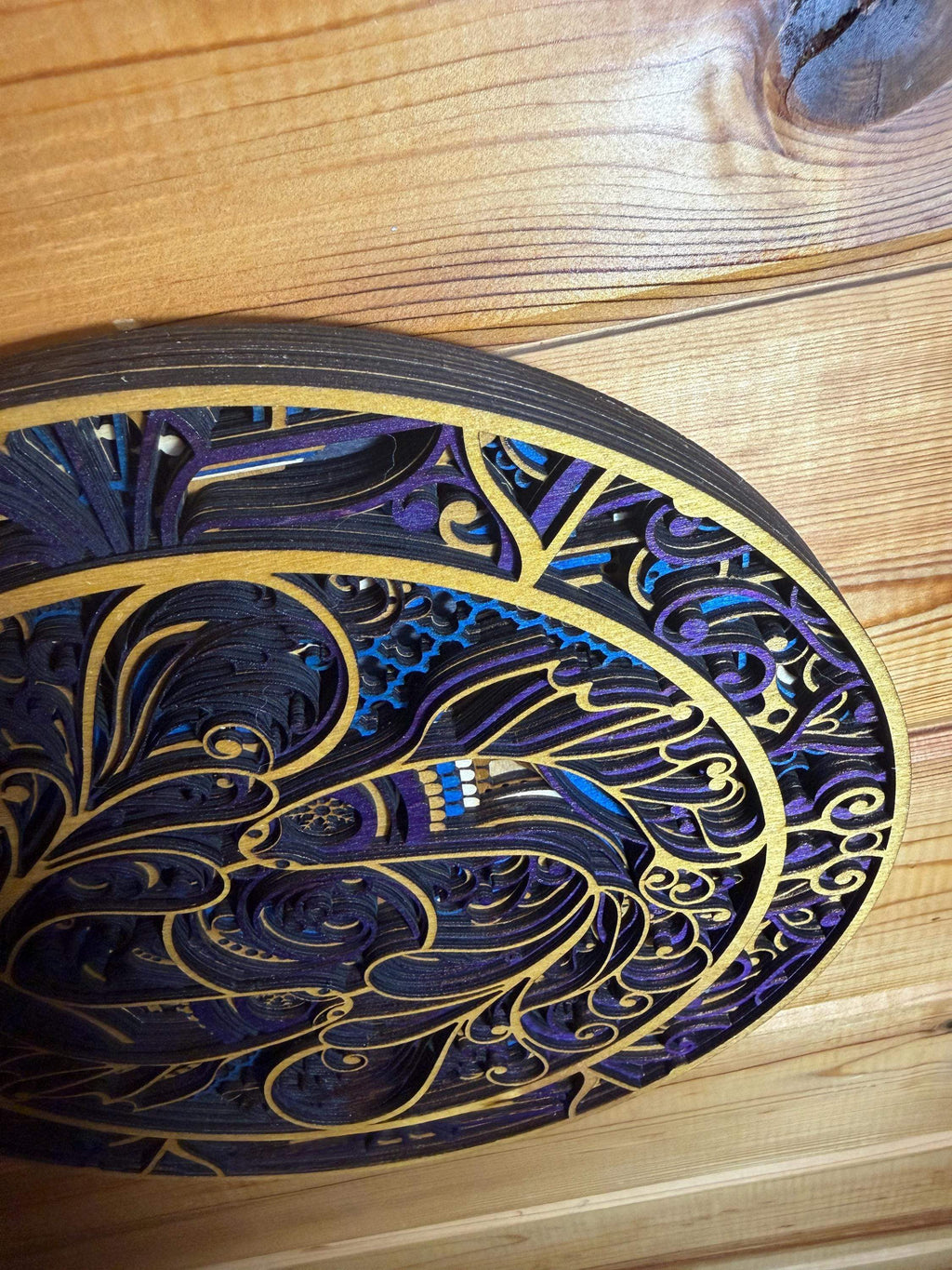 Layered wood mandala with intricate purple, blue, and gold designs on a wooden surface