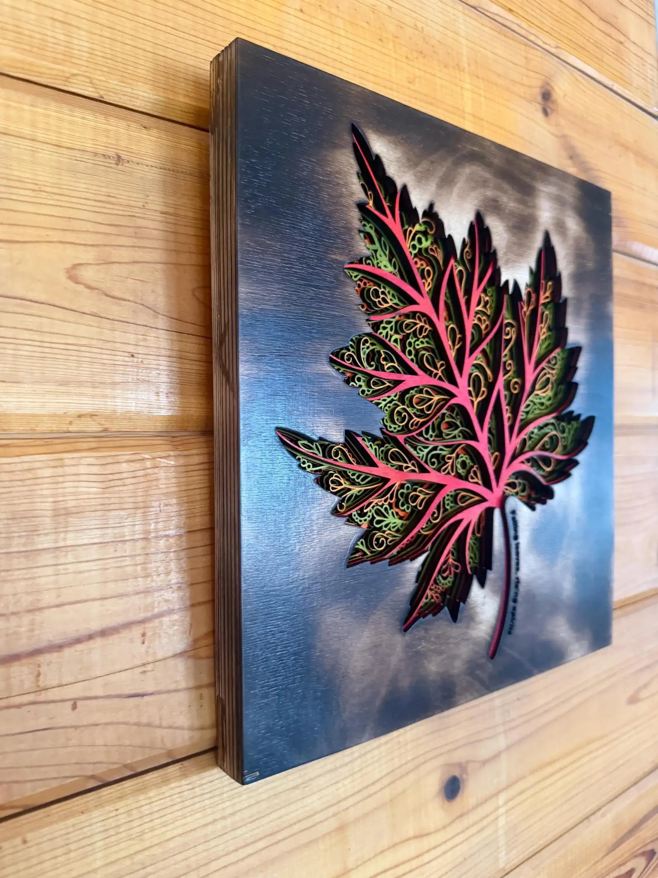Layered wood art maple leaf with intricate green and pink designs on a gradient background