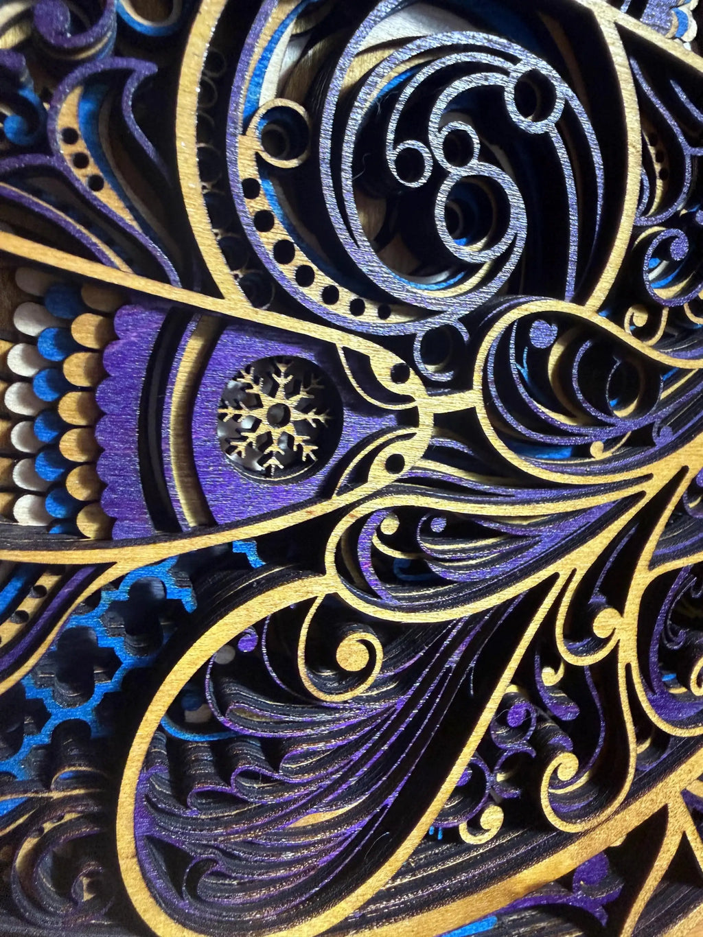 Intricate layered wood art with purple, blue, and gold swirls, featuring a snowflake cutout and detailed laser-cut patterns.
