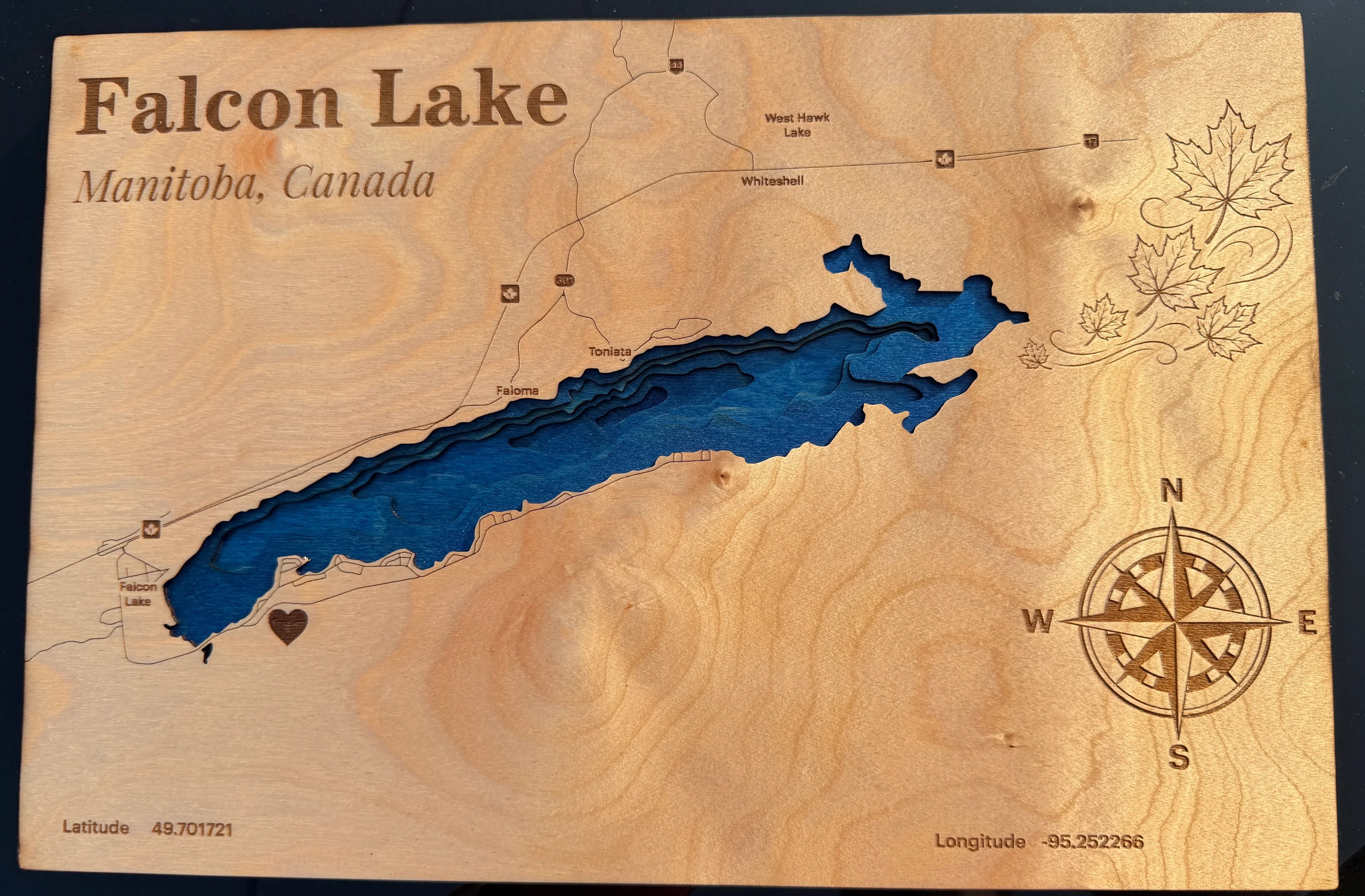 Layered wood map of Falcon Lake, Manitoba, Canada with blue depth layers, compass rose, and maple leaf design.