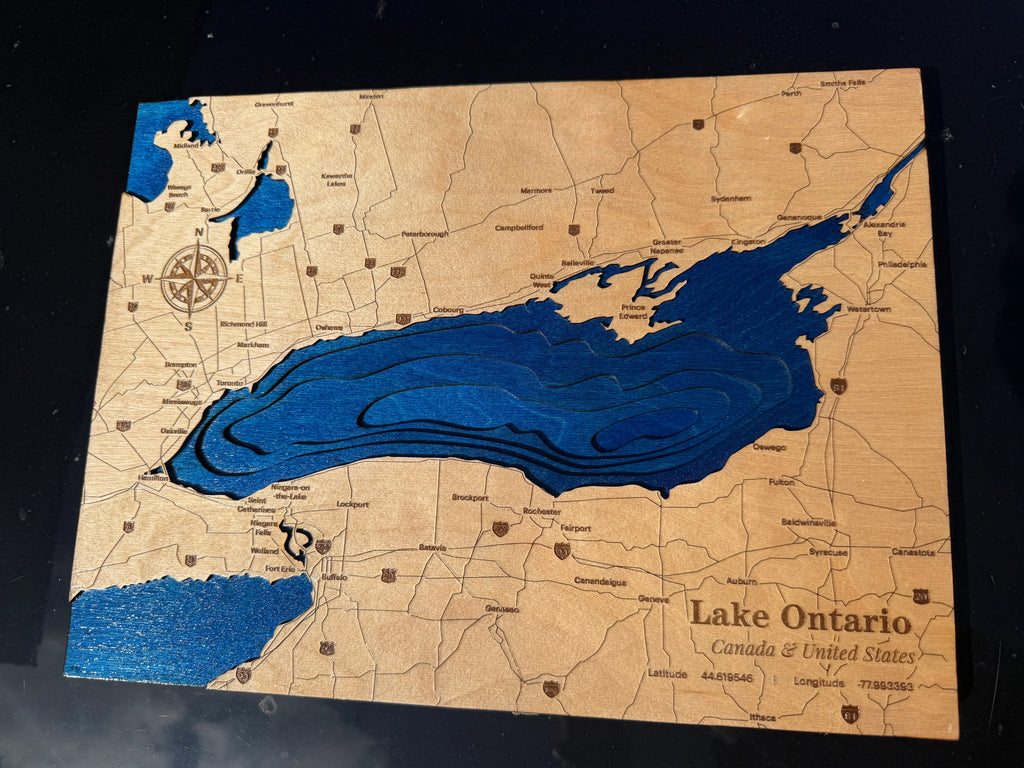 A rectangular wooden map plaque featuring Lake Ontario in deep blue with engraved land details and labels, including 'Lake Ontario Canada & United States' and coordinates, photographed on a dark background.