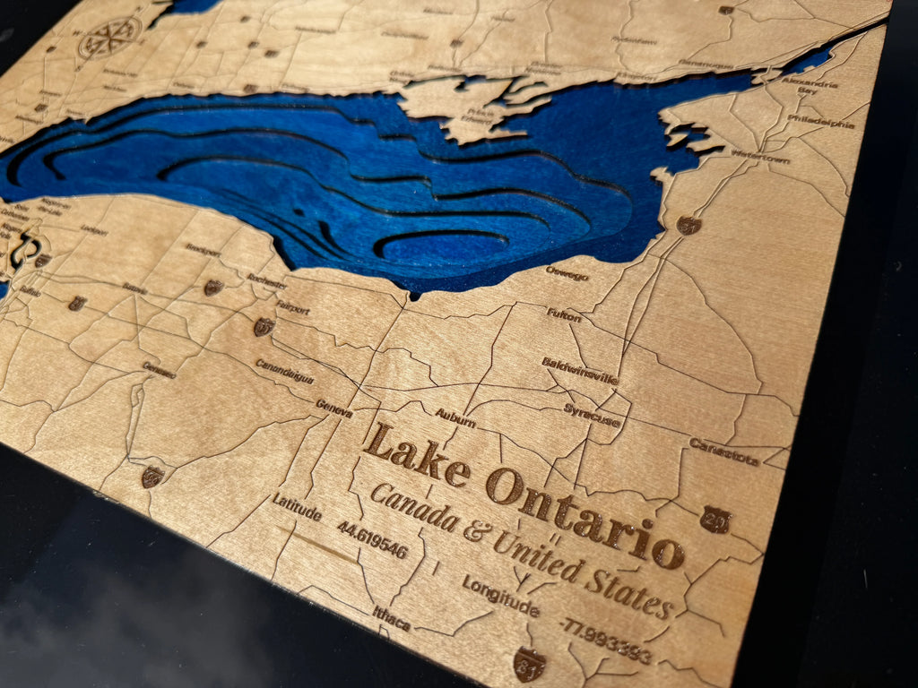 A close-up of a custom wooden map plaque featuring Lake Ontario, with laser-engraved topography lines and text reading 'Lake Ontario Canada & United States' on a natural wood background.