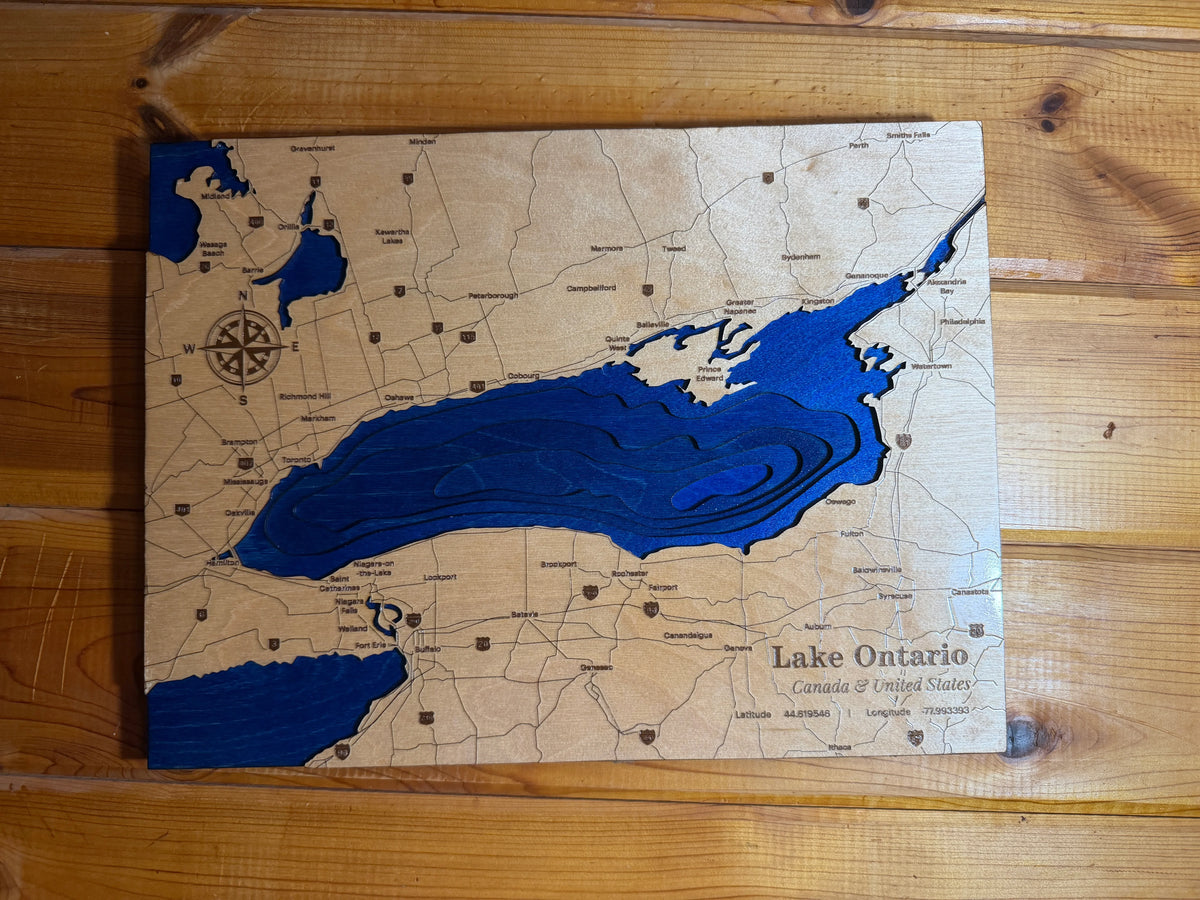 A wooden map plaque of Lake Ontario, featuring a natural wood grain background with laser-etched roads and labels, filled with a vibrant blue resin layer representing the lake. The plaque is photographed from a top-down perspective on a light-colored wooden surface.