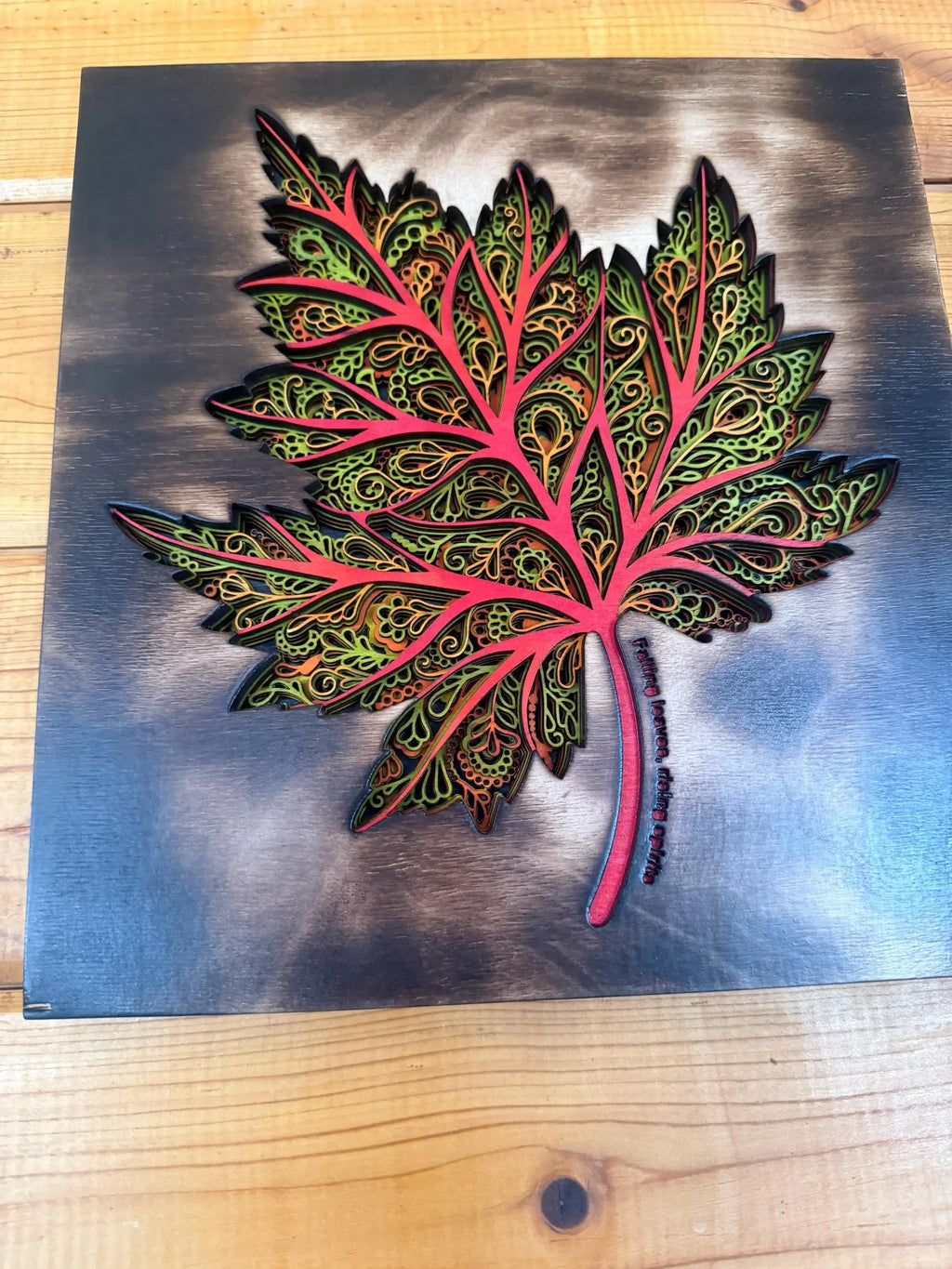 Layered wood art maple leaf with intricate mandala patterns, vibrant red and green hues on dark wood background