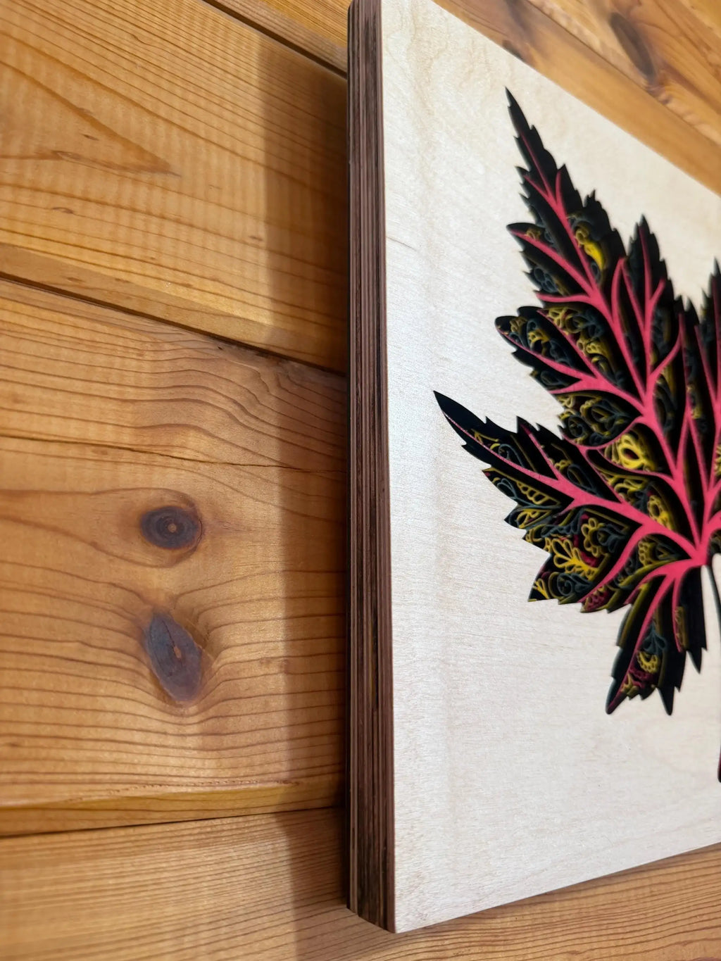 Layered wood art featuring a vibrant, intricately cut leaf design with pink, yellow, and black details on natural wood.