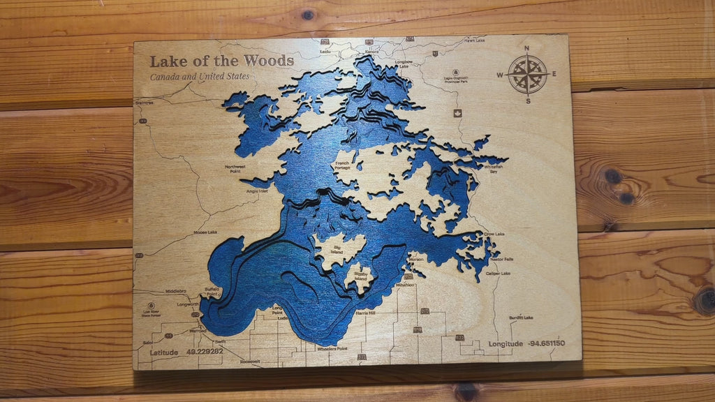 Slow-moving video showcasing a handcrafted layered wooden depth map of Lake of the Woods. The camera pans around the artwork from multiple angles, highlighting the blue wood-dyed water layers, engraved depth contours, detailed shoreline, and laser-etched map features. Close-up views reveal the natural wood grain, engraved compass rose, and the 'Lake of the Woods – Canada and United States' title, giving a clear look at the craftsmanship and dimensional detail of the piece.
