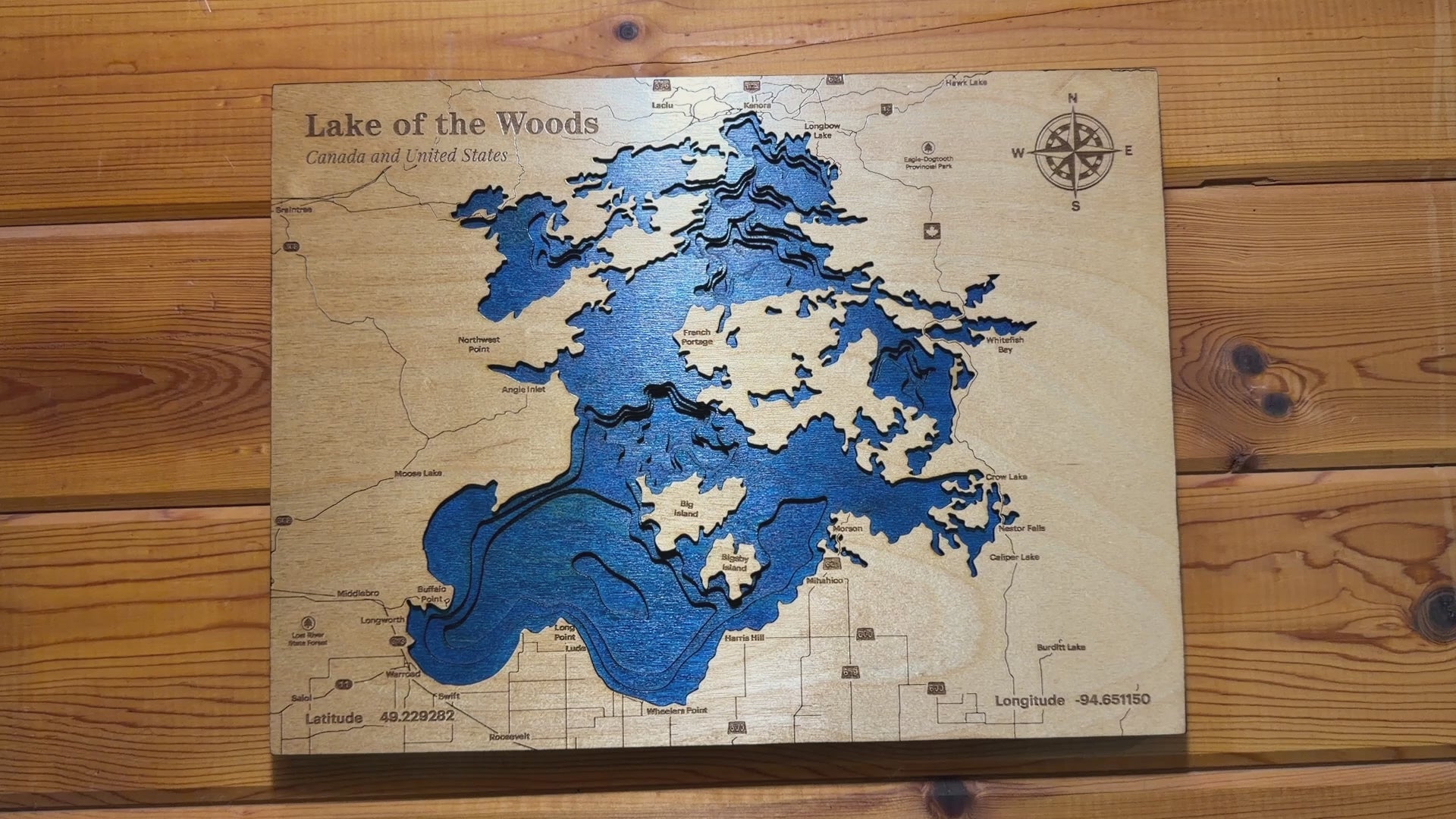 Slow-moving video showcasing a handcrafted layered wooden depth map of Lake of the Woods. The camera pans around the artwork from multiple angles, highlighting the blue wood-dyed water layers, engraved depth contours, detailed shoreline, and laser-etched map features. Close-up views reveal the natural wood grain, engraved compass rose, and the 'Lake of the Woods – Canada and United States' title, giving a clear look at the craftsmanship and dimensional detail of the piece.